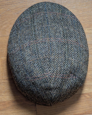 Die Draufsicht zeigt eine Schiebermütze aus Harris Tweed in Detailansicht in den Farben beige und grau, die das Fischgrätmuster bilden. Zusätzlich sind beim Weben feine Fäden in orange und rot eingearbeitet worden, um ein dezentes Strukturkaro zu bilden.