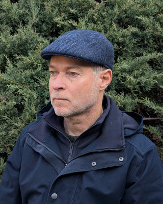 Die Harris Tweed Schiebermütze ist getragen und in Dreiviertelansicht zu sehen. Der Wolltweed ist in einem klassischen Design, nämlich in einem Fischgrätmuster, ausgeführt. Besonders gut ist die Abwechslung der blauen mit schwarzblauen Kette- und Schussfäden.
