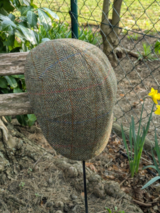 Zu sehen ist eine Schiebermütze aus Harris Tweed in hellem grünem Fischgrätmuster. Außerdem sind zusätzliche Einträge in Kett- und Schussrichtung vorgenommen, die in Rot, Gelb und Blau zu erkennen sind.