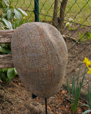 Zu sehen ist eine Schiebermütze aus Harris Tweed in einem freundlichen Sandtonfarbe. Der Tweed ist als Fischgrät gestaltet. Außerdem sind zusätzliche Kett- und Schussfäden in anderen Farben eingetragen, die ein Musterkaro bilden.